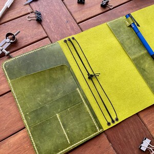 K&ouml;nnte beinhalten: Offenes Lederjournal mit limettengr&uuml;nem Innenraum und olivgr&uuml;nen Taschen. Das Journal ist ge&ouml;ffnet und zeigt einen Stift und einen elastischen Verschluss. Werkzeuge wie ein Lineal, B&uuml;roklammern und ein Bastelmesser liegen auf der Holzoberfl&auml;che.
