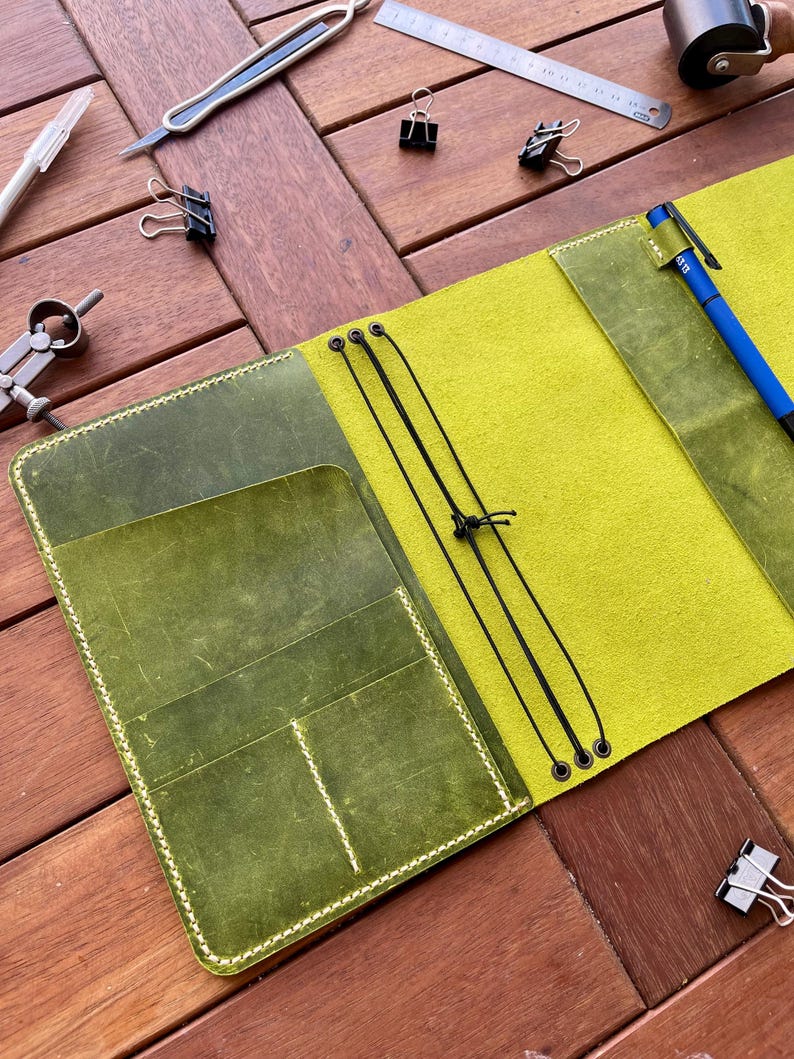 K&ouml;nnte beinhalten: Offenes Lederjournal mit limettengr&uuml;nem Innenraum und olivgr&uuml;nen Taschen. Das Journal ist ge&ouml;ffnet und zeigt einen Stift und einen elastischen Verschluss. Werkzeuge wie ein Lineal, B&uuml;roklammern und ein Bastelmesser liegen auf der Holzoberfl&auml;che.