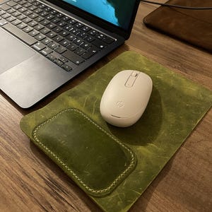May include: A rectangular olive green leather mouse pad with a stitched wrist rest, featuring a distressed texture. A white HP wireless computer mouse sits on the surface of the pad, positioned next to a dark gray laptop with a black keyboard.