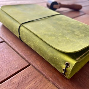 K&ouml;nnte beinhalten: Ein gr&uuml;nes Lederjournal mit Klappenverschluss und schwarzem Gummiband. Das Journal ist etwa 20 cm lang und 13 cm breit. Ein kleines Rollwerkzeug liegt auf der Holzoberfl&auml;che.