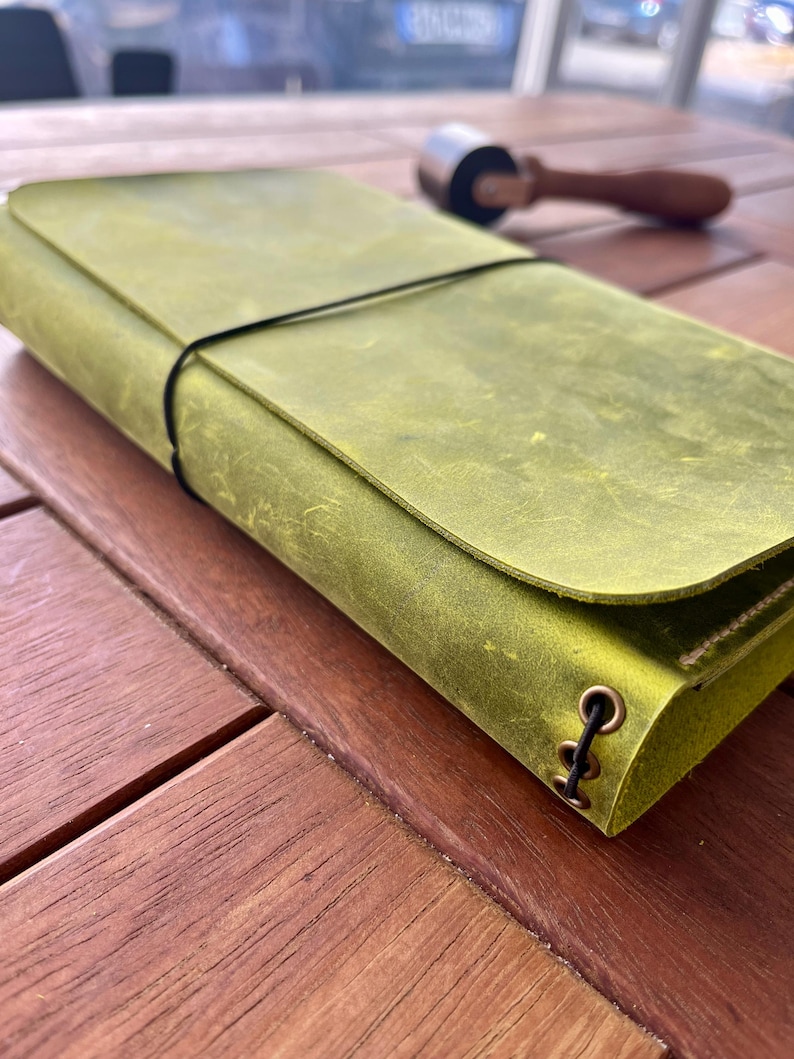 K&ouml;nnte beinhalten: Ein gr&uuml;nes Lederjournal mit Klappenverschluss und schwarzem Gummiband. Das Journal ist etwa 20 cm lang und 13 cm breit. Ein kleines Rollwerkzeug liegt auf der Holzoberfl&auml;che.