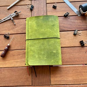 K&ouml;nnte beinhalten: Ein gr&uuml;nes Lederjournal mit schwarzem Gummibandverschluss, umgeben von verschiedenen Schreibwaren, darunter ein Stift, ein Lineal und B&uuml;roklammern. Das Journal hat eine strukturierte Oberfl&auml;che und ein rotes Band als Lesezeichen. Auf einer Holzoberfl&auml;che.
