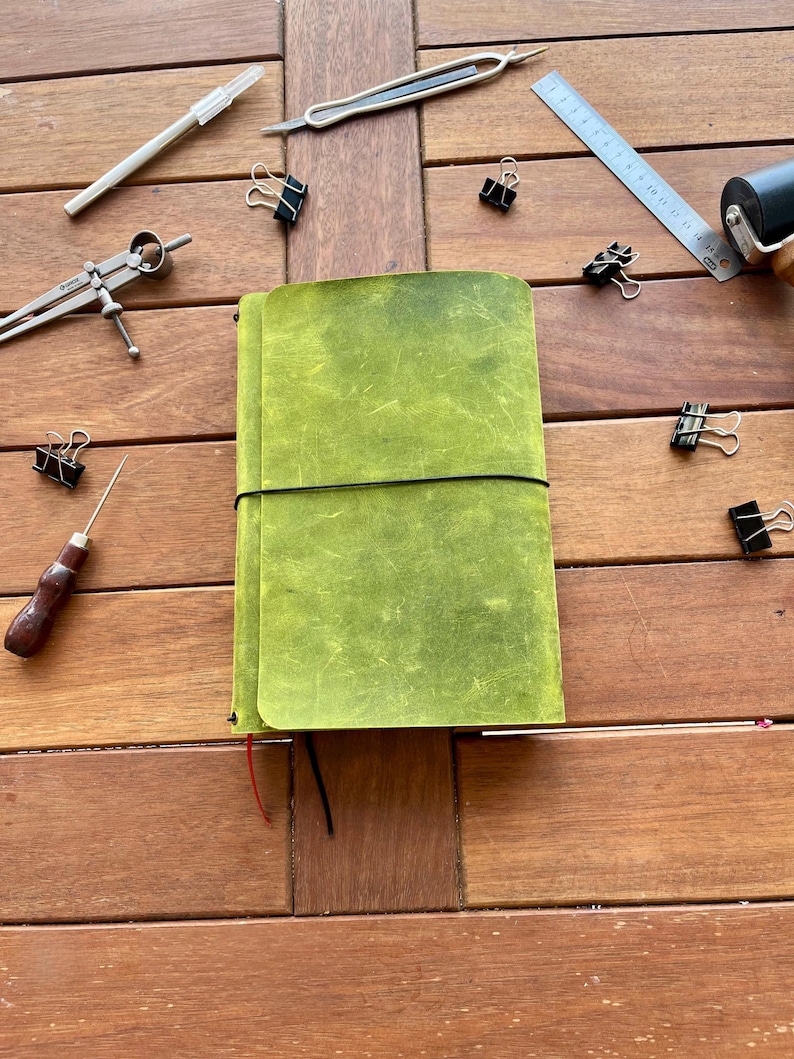K&ouml;nnte beinhalten: Ein gr&uuml;nes Lederjournal mit schwarzem Gummibandverschluss, umgeben von verschiedenen Schreibwaren, darunter ein Stift, ein Lineal und B&uuml;roklammern. Das Journal hat eine strukturierte Oberfl&auml;che und ein rotes Band als Lesezeichen. Auf einer Holzoberfl&auml;che.