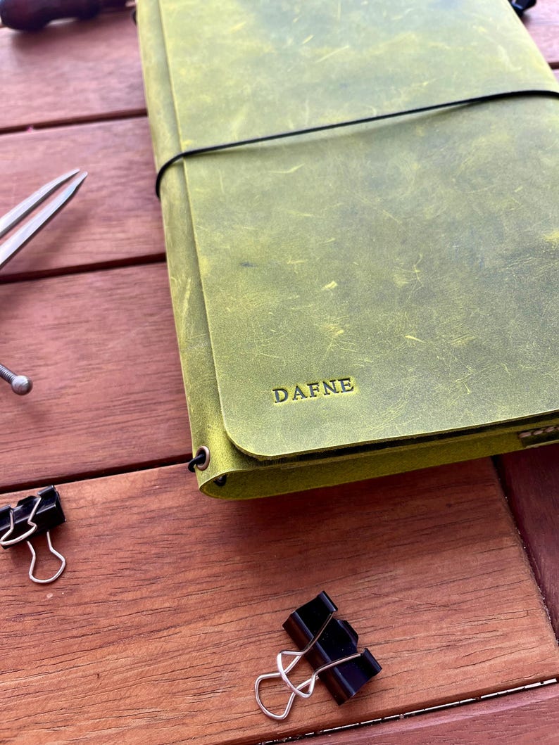 K&ouml;nnte beinhalten: Ein gr&uuml;nes Lederjournal mit dem Namen "DAFNE" auf dem Einband. Das Journal wird mit einem schwarzen Gummiband verschlossen. Zwei schwarze B&uuml;roklammern und eine silberne Schere sind ebenfalls zu sehen.