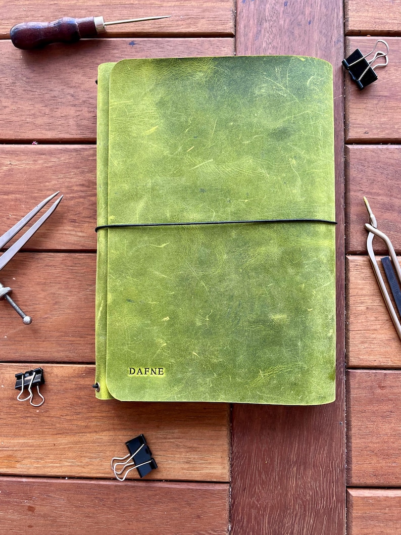 K&ouml;nnte beinhalten: Ein gr&uuml;nes Lederjournal mit dem Namen "DAFNE" auf dem Einband. Das Journal wird mit einem schwarzen Gummiband verschlossen. Verschiedene Bastelwerkzeuge und B&uuml;roklammern sind um das Journal herum auf einer Holzoberfl&auml;che angeordnet.