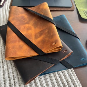 May include: Three leather notebook covers in tan, dark brown, and blue, each secured with a black elastic band. The covers feature a smooth, matte texture and are embossed with the initials TJD in the bottom right corner of each piece.