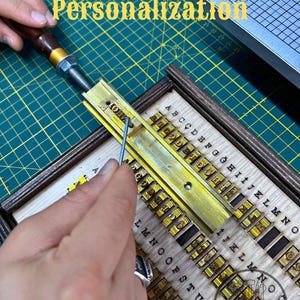 K&ouml;nnte beinhalten: Nahaufnahme eines Metallpr&auml;gewerkzeugs mit Holzgriff und Messing-Schrifttr&auml;ger. Der Text "Personalisierung" ist gelb dargestellt. Das Werkzeug ist mit einzelnen Buchstabenstempeln ausgestattet.