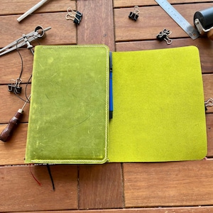K&ouml;nnte beinhalten: Offenes gr&uuml;nes Lederjournal mit hellgr&uuml;nem Innenraum, blauem Stift und rotem Lesezeichen. Verschiedene Schreibwaren, darunter ein Lineal, B&uuml;roklammern und eine Rolle, sind um das Journal auf einer Holzoberfl&auml;che angeordnet.