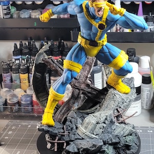Może przedstawiać: Szczegółowa figurka postaci Cyclops z X-Men w dynamicznej pozie. Figurka jest głównie niebieska i żółta, ze szczegółową twarzą i wizjerem. Stoi na skalistej, szarej podstawie, prezentując scenę walki. Figurka ma około 25 cm wysokości.