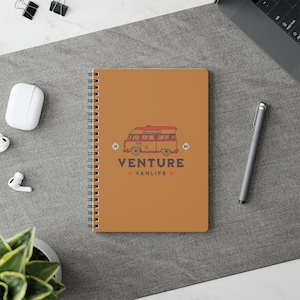 May include: A spiral-bound notebook with a brown cover featuring a red and brown graphic of a vintage van. The text "VENTURE VANLIFE" is below the van illustration. A pen, wireless earbuds, and a laptop are visible.