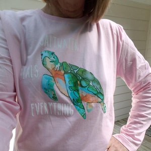 May include: Pink long-sleeved t-shirt with a watercolor illustration of a sea turtle and the text "Saltwater heals everything."