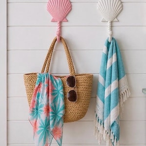 May include: Two wall-mounted hooks shaped like seashells, one pink and one white, each featuring a twisted stem. A woven straw tote bag with a floral scarf and brown sunglasses hangs on the pink hook, while a blue and white striped towel hangs on the white hook.