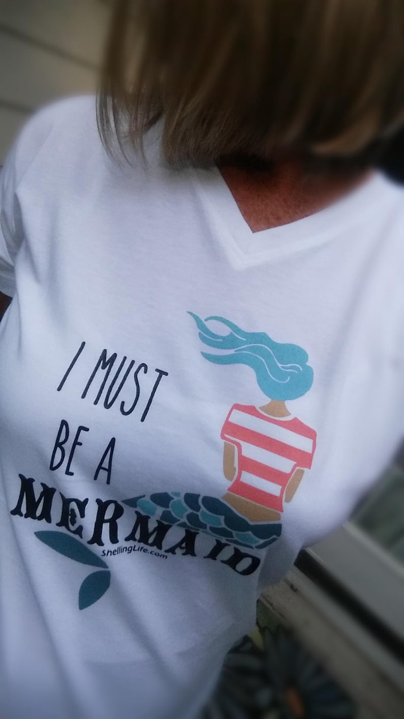 May include: White t-shirt with a black graphic that says "I must be a mermaid" with a mermaid illustration in teal, pink, and white.