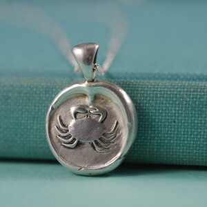 May include: Silver pendant necklace featuring a detailed crab design. The pendant is round and has a textured surface. The necklace is on a teal fabric background.