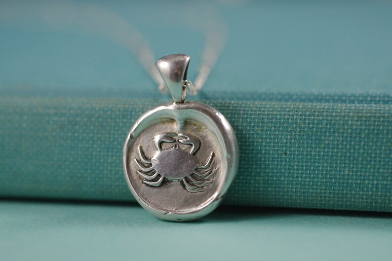 May include: Silver pendant necklace featuring a detailed crab design. The pendant is round and has a textured surface. The necklace is on a teal fabric background.