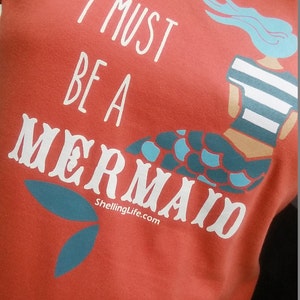 May include: Red t-shirt with a white graphic of a mermaid with the text "I must be a mermaid" and the website "ShellingLife.com".