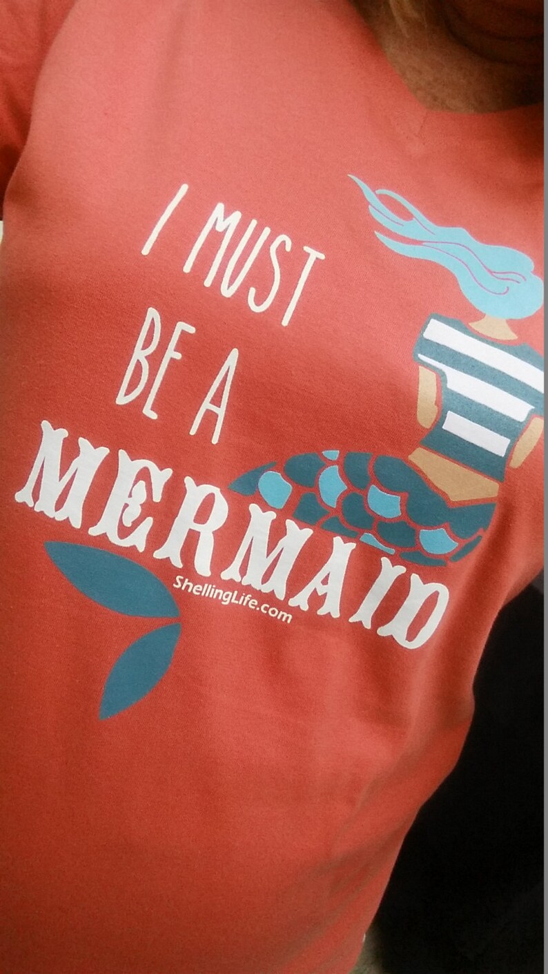 May include: Red t-shirt with a white graphic of a mermaid with the text "I must be a mermaid" and the website "ShellingLife.com".