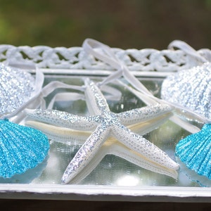 May include: Four decorative seashells, two blue and two silver, with a glittery white starfish on a white tray.