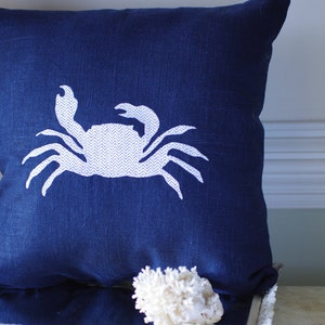 May include: A navy blue throw pillow with a white embroidered crab design. The crab is facing right and has a chevron pattern on its body.