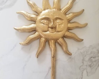 Op de afbeelding: Gouden metalen zonvormige wandhaak met een glimlachend gezicht. De haak is bevestigd aan de onderkant van de zon.