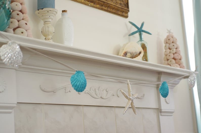 May include: A white mantelpiece decorated with a seashell garland, starfish, and a seashell Christmas tree. The garland is made of blue seashells and white string.