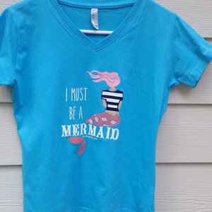 May include: A turquoise blue v-neck t-shirt with a graphic of a mermaid with pink hair and a striped shirt. The text "I must be a mermaid" is printed on the shirt.