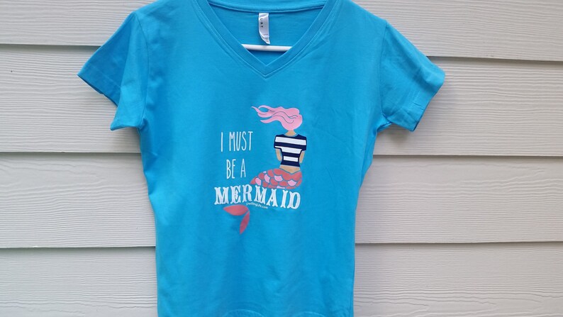 May include: A turquoise blue v-neck t-shirt with a graphic of a mermaid with pink hair and a striped shirt. The text "I must be a mermaid" is printed on the shirt.
