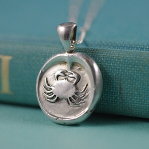 May include: Silver pendant necklace featuring a detailed crab design. The pendant is round and has a textured surface. The necklace is on a blue book.