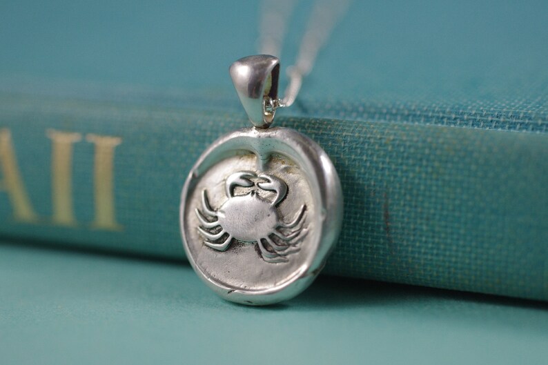 May include: Silver pendant necklace featuring a detailed crab design. The pendant is round and has a textured surface. The necklace is on a blue book.
