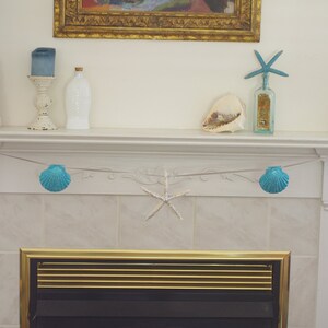 May include: A white fireplace mantel decorated with a seashell garland, two pink seashell towers, a blue starfish, and a blue seashell garland. The fireplace has a black grate with a gold trim.