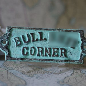May include: A teal blue metal sign with the words "BULL CORNER" in black lettering. The sign has a distressed, rustic look.