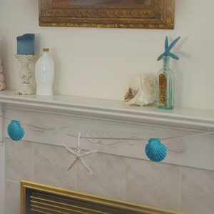 May include: A white mantelpiece with a seashell garland, two pink seashell trees, a blue glass bottle with a starfish on top, a white bottle, a white candle holder with a blue candle, and a seashell.