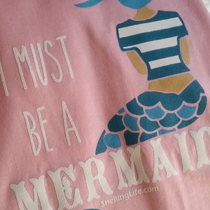 May include: Pink t-shirt with a white graphic of a mermaid with the text "I must be a mermaid" and the website "ShellingLife.com".