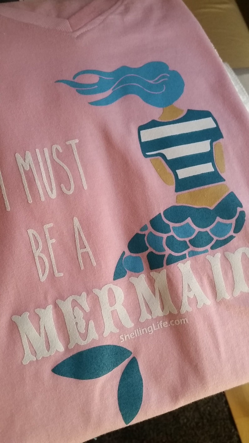 May include: Pink t-shirt with a white graphic of a mermaid with the text "I must be a mermaid" and the website "ShellingLife.com".