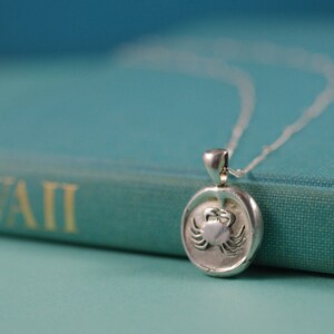 May include: A silver necklace with a round pendant featuring a detailed crab design. The pendant is resting on a teal book with the word "HAWAII" printed on the cover.