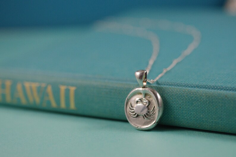 May include: A silver necklace with a round pendant featuring a detailed crab design. The pendant is resting on a teal book with the word "HAWAII" printed on the cover.