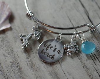 May include: A silver charm bracelet with multiple charms, including a charm with the text "life's a beach", a seashell charm, a crab charm, and a blue gemstone charm.