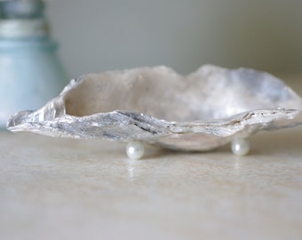 Puede incluir: Un plato de concha de ostra plateado con una perla a cada lado, utilizada como patas para elevar el plato.