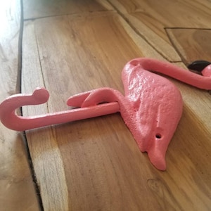May include: A pink flamingo-shaped wall hook made of cast iron. The hook is painted in a matte pink color with a black beak. The flamingo is lying on a wooden surface.