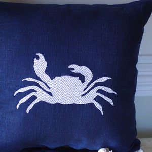 May include: A navy blue throw pillow with a white embroidered crab design. The crab is facing right and has a chevron pattern on its body.