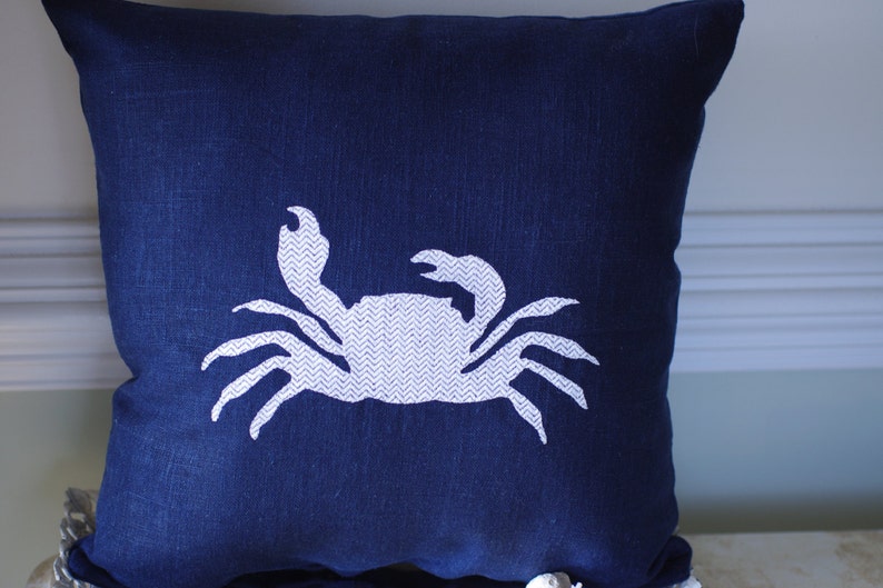 May include: A navy blue throw pillow with a white embroidered crab design. The crab is facing right and has a chevron pattern on its body.