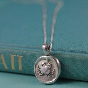 May include: A silver necklace with a round pendant featuring a detailed crab design. The pendant is on a blue book with the word "All" visible.