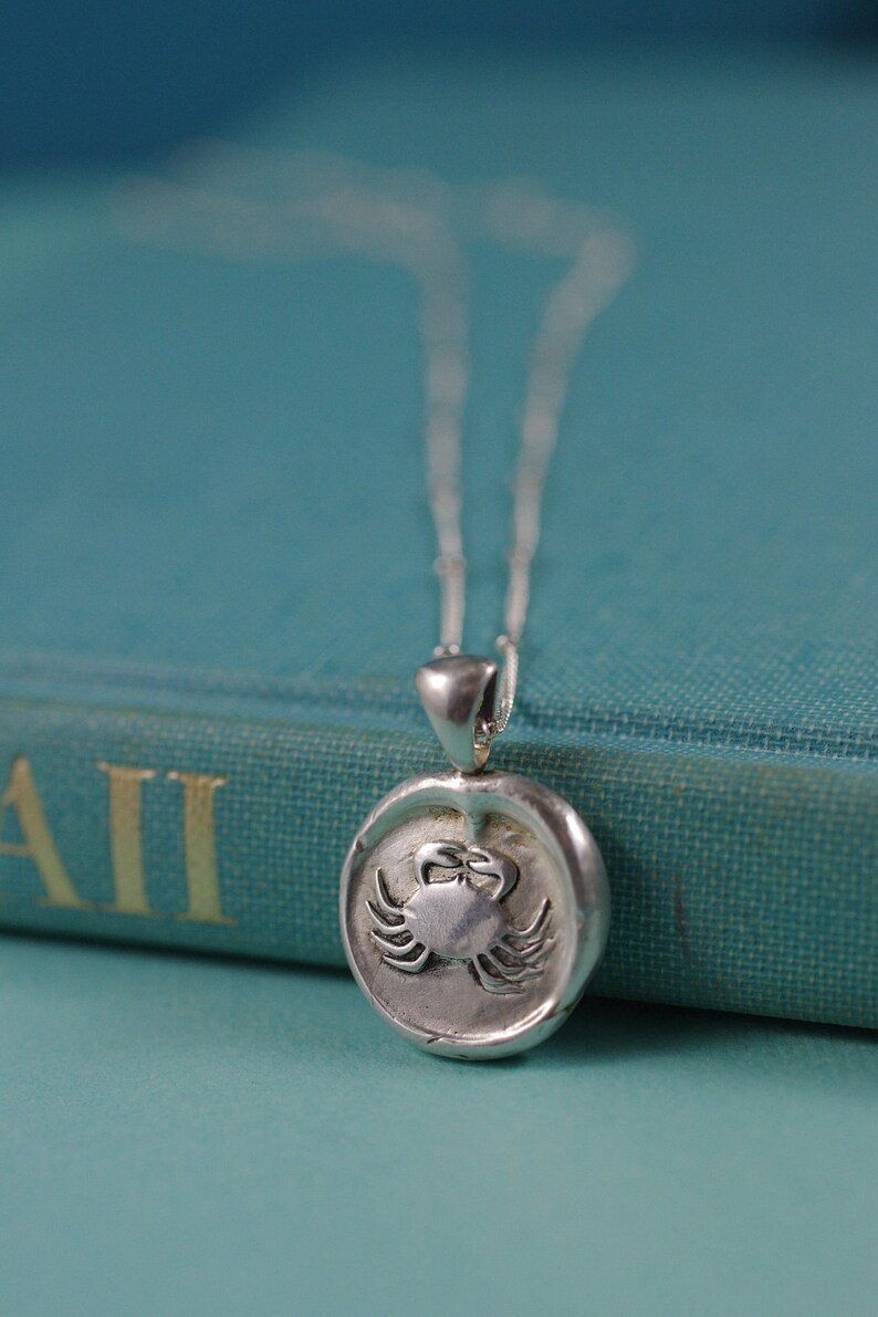 May include: A silver necklace with a round pendant featuring a detailed crab design. The pendant is on a blue book with the word "All" visible.