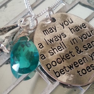 May include: Silver necklace with a teal gemstone, a seashell charm, and a silver oval pendant with the inscription "may you always have a shell in your pocket & sand between your toes".