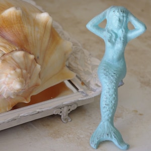 May include: A light blue mermaid figurine with a seashell in a white tray. The mermaid is sitting with her arms raised above her head.