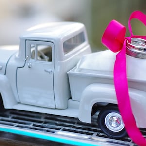 May include: A white toy pickup truck with a pink ribbon and two silver wedding bands on top. The truck is on a glass surface.