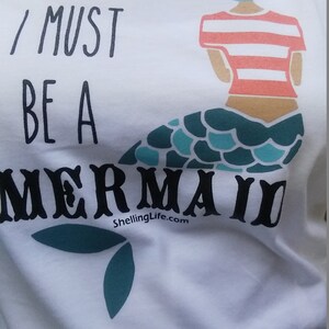 May include: White t-shirt with a blue and green mermaid graphic and the text "I must be a mermaid" and "ShellingLife.com".