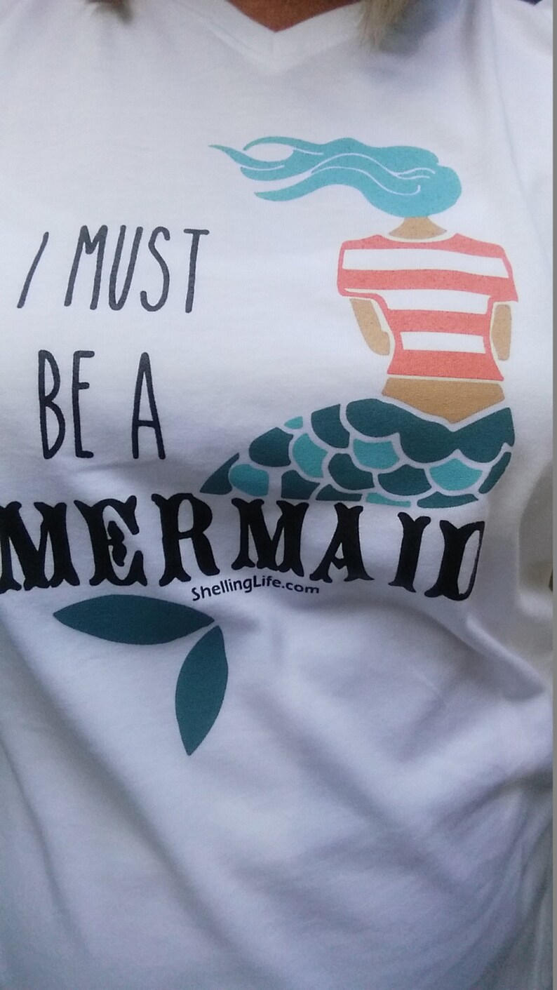 May include: White t-shirt with a blue and green mermaid graphic and the text "I must be a mermaid" and "ShellingLife.com".