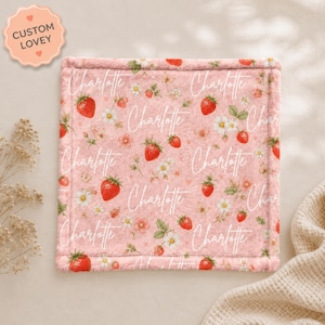 Può includere: Una copertina di sicurezza quadrata in morbido tessuto rosa con un motivo ripetuto di fragole rosse, margherite bianche e piccoli fiori rosa. Il nome Charlotte è stampato in corsivo bianco ripetutamente sul tessuto.