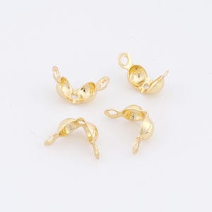 May include: Four small, gold-colored crimp bead findings, used in jewelry making. Each bead is open, showing the interior, with small loops for attaching to other components. The beads are scattered on a white background.